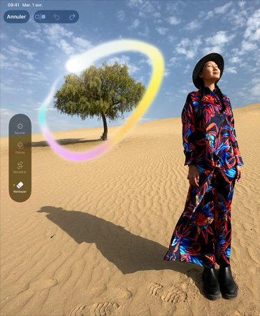 Photo d’une femme dans une robe multicolore debout dans un paysage désertique, à l’arrière-plan un arbre est encerclé d’un arc-en-ciel lumineux pour illustrer la capacité d’Apple Intelligence à supprimer les objets en arrière-plan