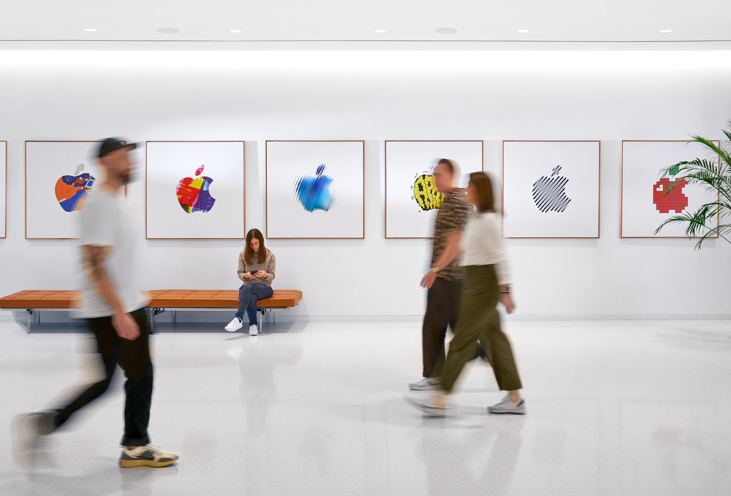 Trois membre de l’équipe Trois membres de l’équipe Apple passent devant un mur orné de logos Apple colorés, et une employée est assise sur un banc.