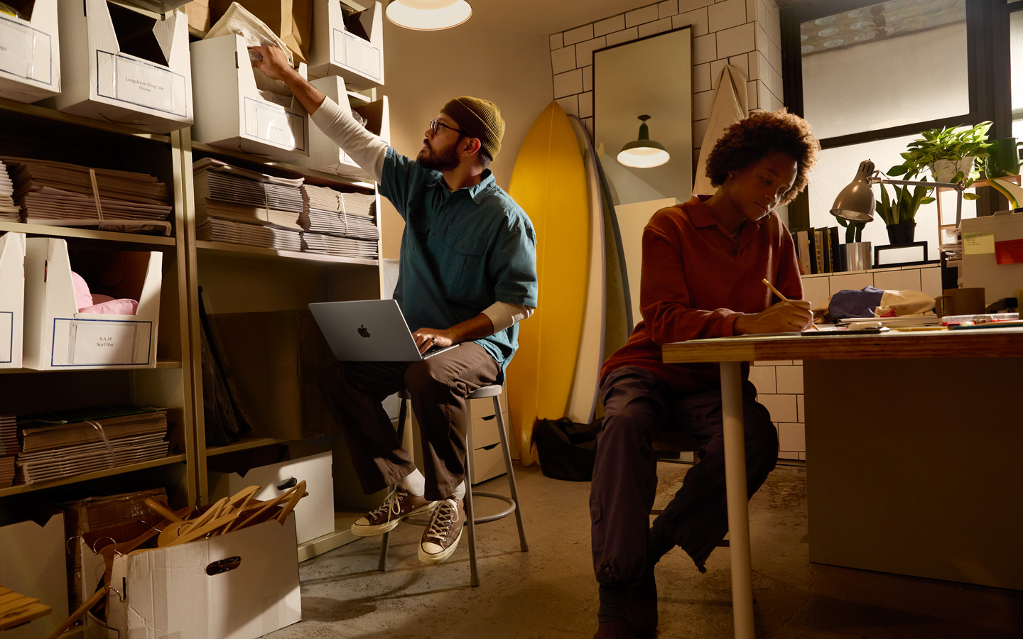 A person sitting at a desk uses a pencil to write on a notebook, another person sitting by a shelf has a MacBook on their lap and reaches for a file with their right hand.