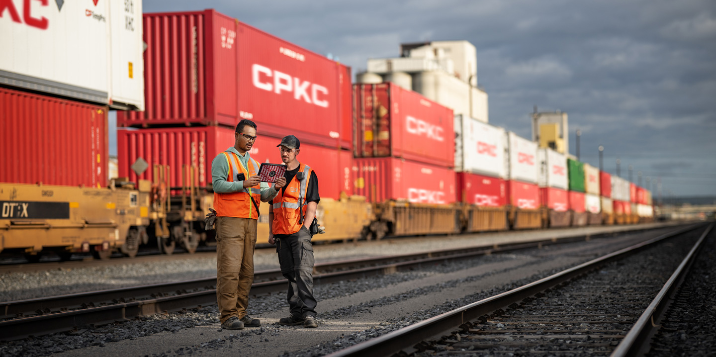To jernbanearbejdere ser på en iPad, mens de står på et baneområde ved et holdende tog med skibscontainere