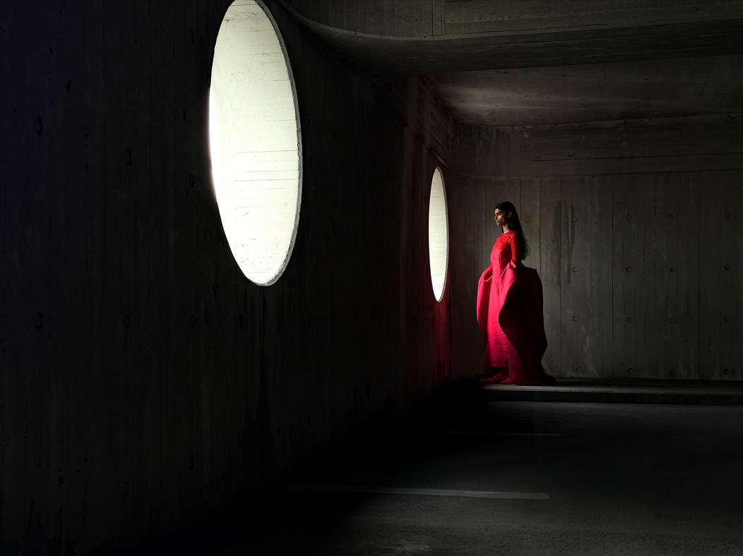 A woman wearing a long red dress stands in an empty room being photographed. The Photo app toggles through the following focal lengths to demonstrate the capabilities of the pro camera system: 200 mm, 100 mm, 48 mm, 35 mm, 28 mm, 13 mm, and macro